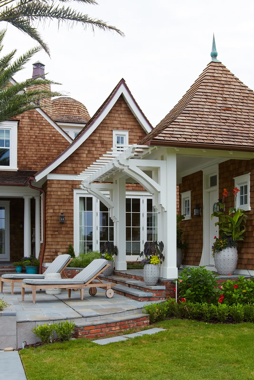Patio with gray lounge chairs, stone floor, potted plants, and large windows with a gabled roof.