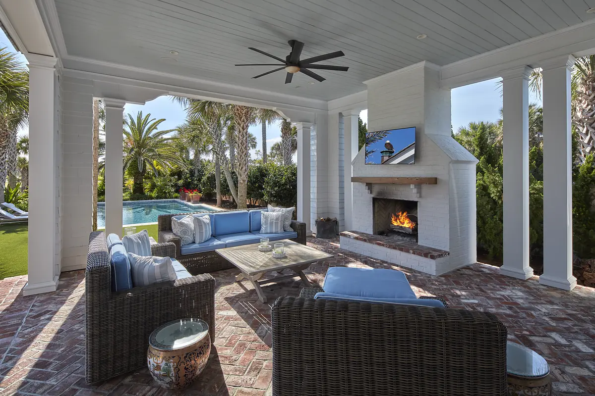 Patio with wicker seating, wooden coffee table, stone fireplace, and mounted TV surrounded by greenery and pool view.