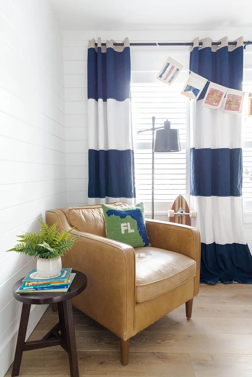 Living room corner with leather armchair, side table, books, potted plant, and blue and white curtains.