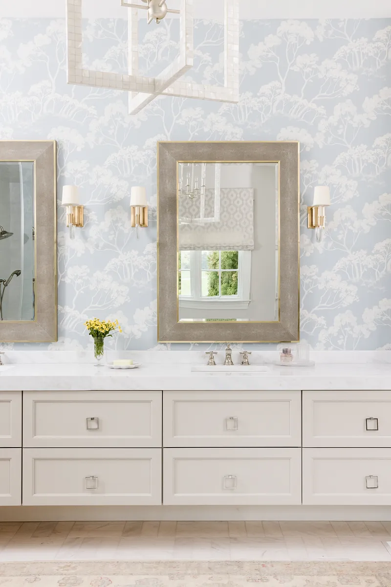 Bathroom with double vanity, mirrors, sconces, and light blue floral wallpaper.