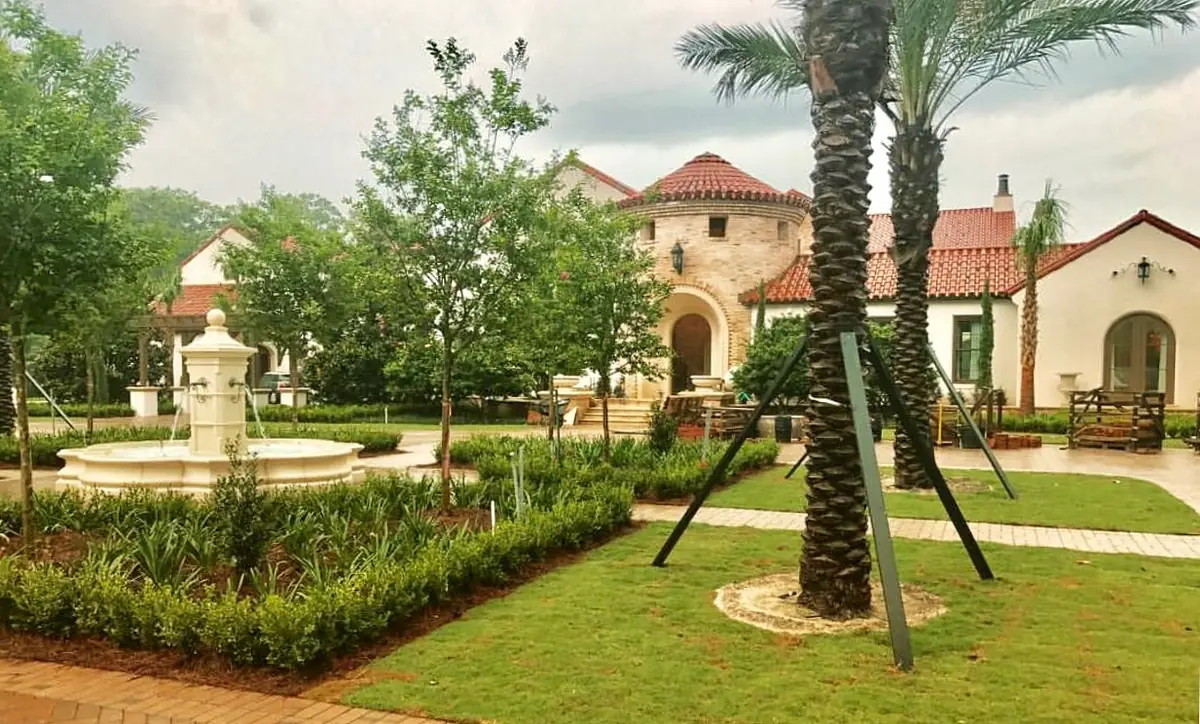 Garden with central fountain, shrubs, palm trees, and brick pathways alongside a building with a red-tiled roof.