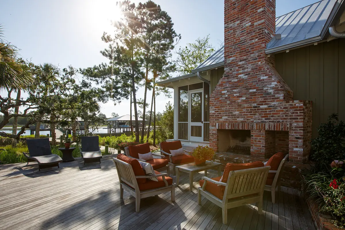 Outdoor deck with wooden floor, brick fireplace, seating, coffee table, and greenery.