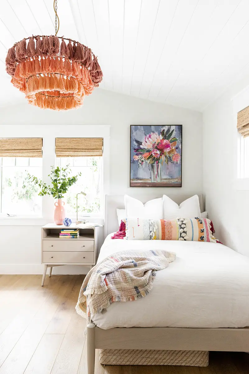 Bedroom with white bed, colorful pillows, nightstand, floral painting, and greenery in a vase.