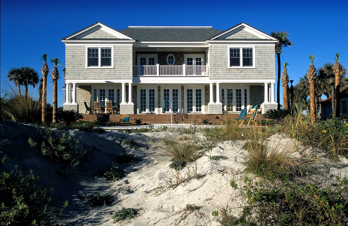 Two-story house with gray shingle exterior, white trim, porch, and palm trees in sandy landscaping.