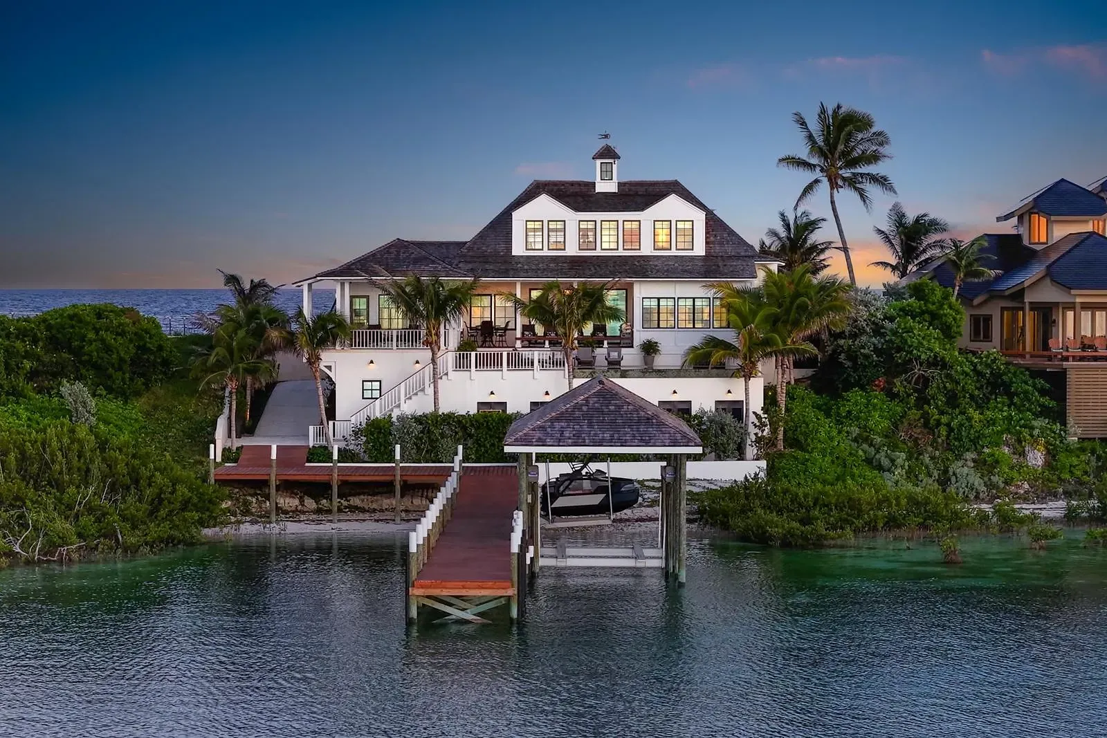 Waterfront house with gabled roof, dock, tropical landscaping, and multiple windows.