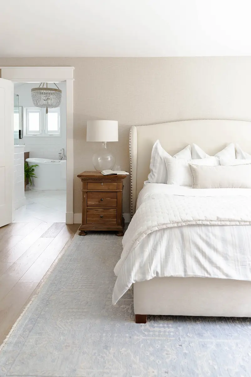 Bedroom with beige upholstered bed, white striped quilt, wooden nightstand, and light blue area rug.