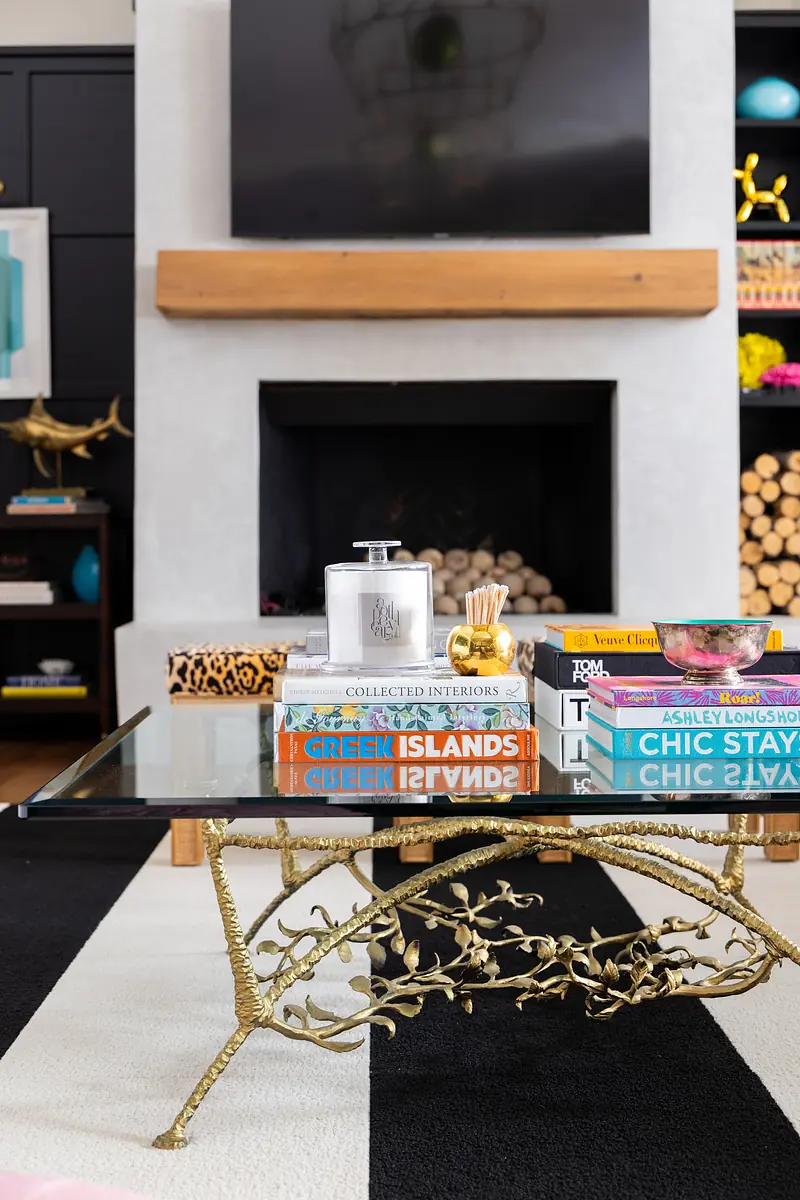 Living room with glass coffee table, stacked books, decorative items, and a textured fireplace wall with art.