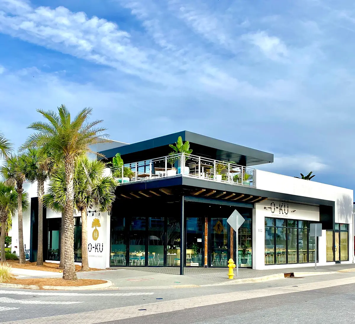 Exterior view of a modern building with glass windows, palm trees, and roof terrace labeled 'O-KU'.