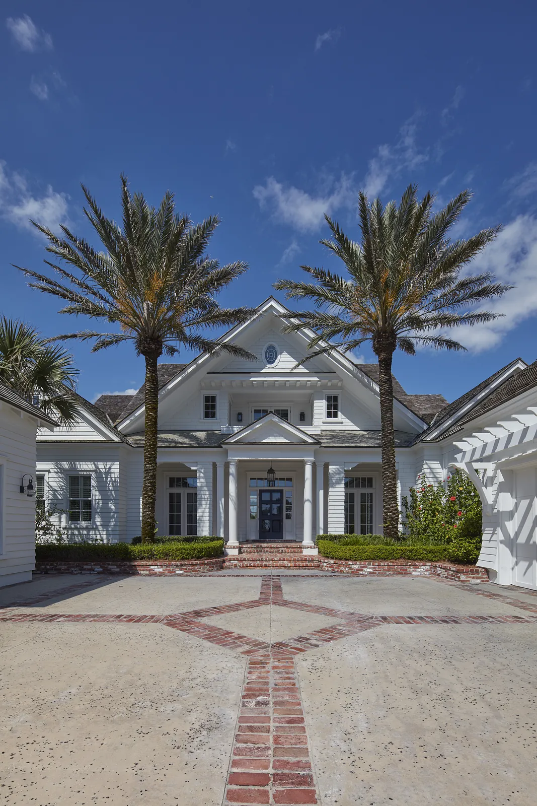 Front exterior of a house with palm trees, brick driveway, and a central entrance with a gable roof.