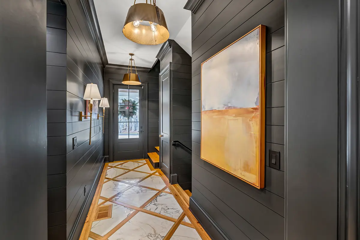 Hallway with dark paneled walls, marble floor, abstract painting, wall sconces, and stairs.