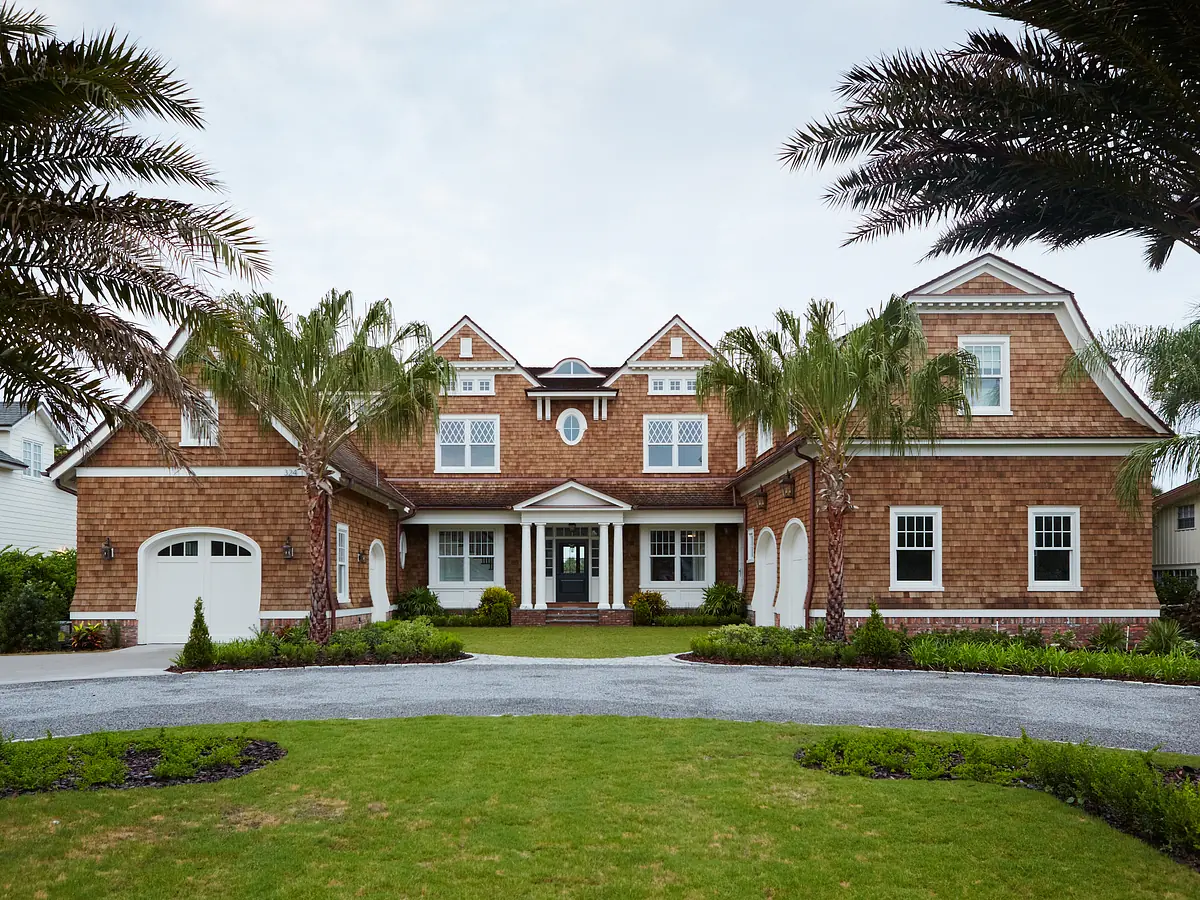 Front exterior of a house with shingle facade, gables, large windows, gravel driveway, and landscaping.