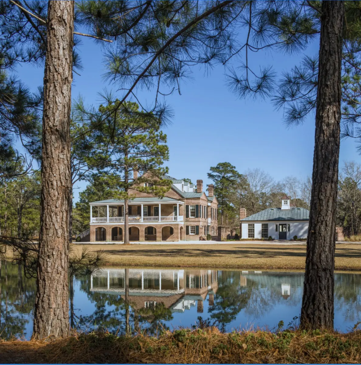 Two-story brick house with large porch, surrounded by trees and a reflective pond.