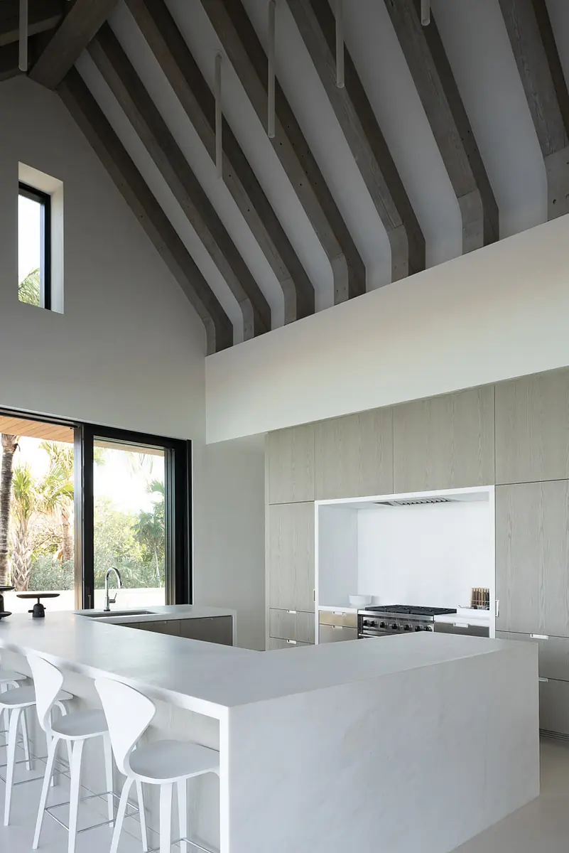 Kitchen with white island, bar stools, wall-mounted range hood, cabinetry, and high ceiling with beams.