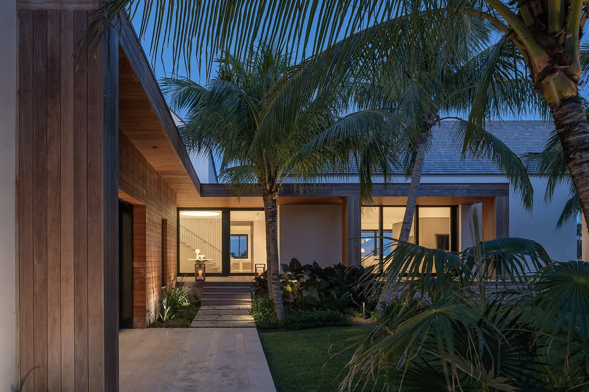 Exterior front view with wood and stone cladding, large windows, palm trees, and a paved stone pathway.