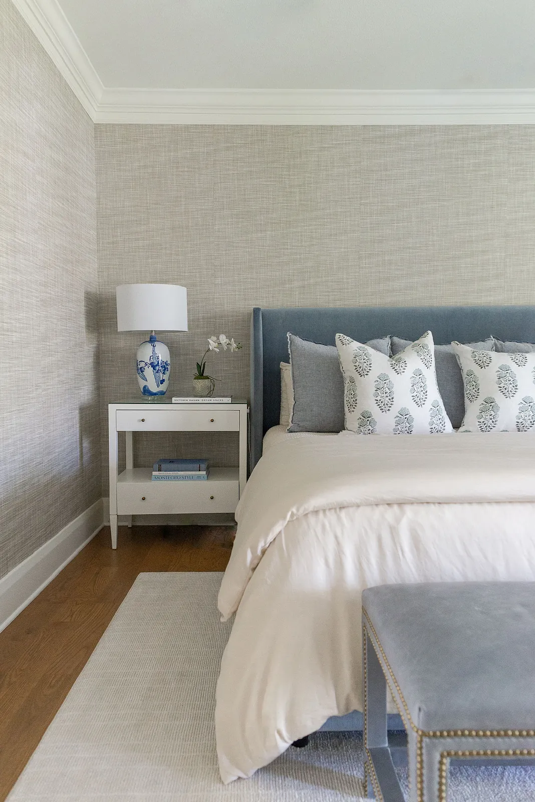 Bedroom with blue bed, gray and white pillows, white nightstand, lamp, small blue ottoman, and beige wallpaper.