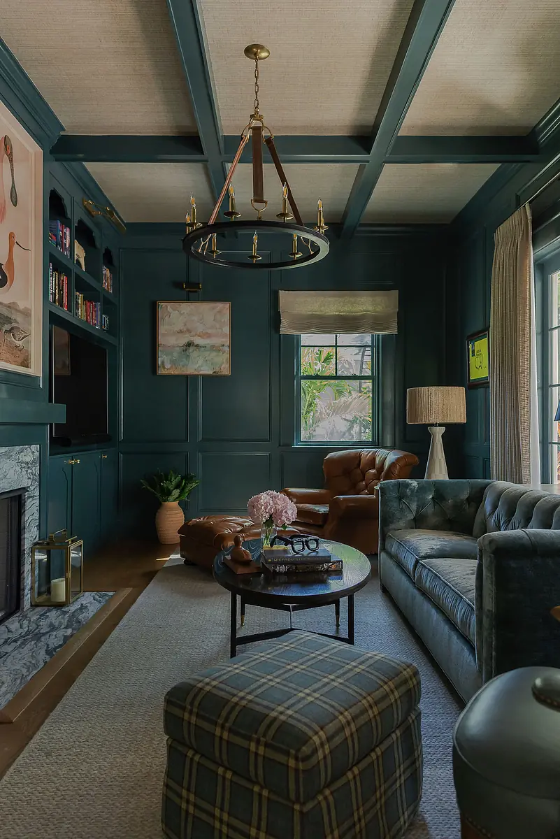 Living room with dark teal walls, tufted green sofa, round coffee table, patterned ottoman, and chandelier
