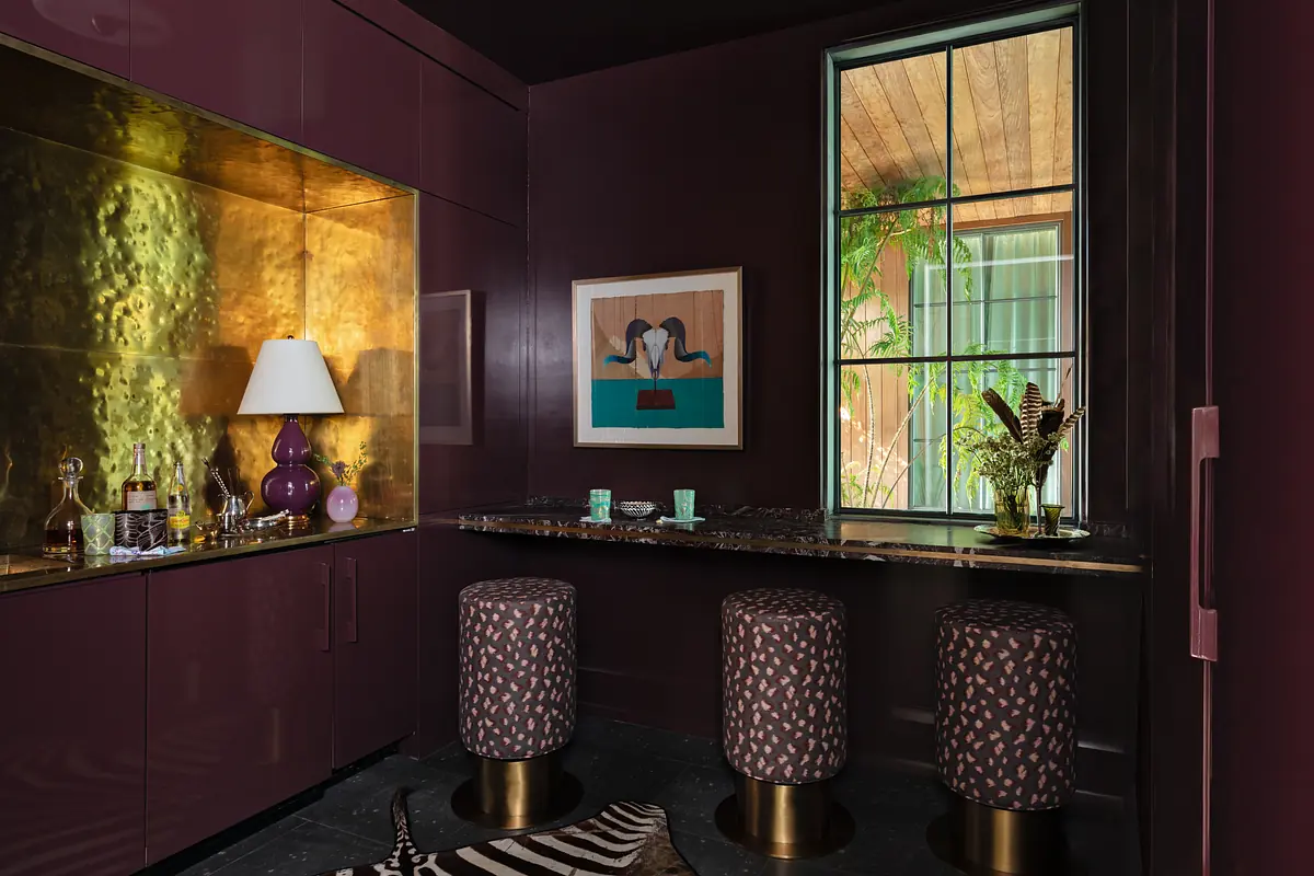 Bar area with purple cabinetry, gold backsplash, granite countertop, stools, artwork, and window with plants.
