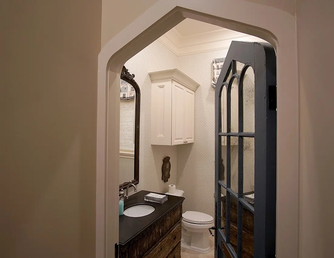 Powder room with pedestal sink, toilet, wooden cabinet, wall mirror, patterned wallpaper, and glass panel door.