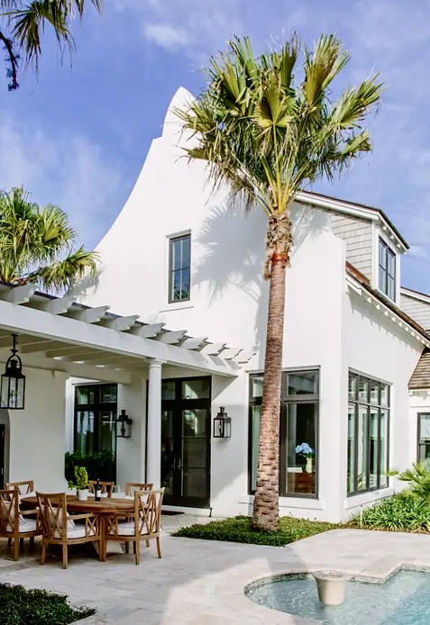 Exterior rear area with dining table, palm trees, pool, large windows, and white stucco house.