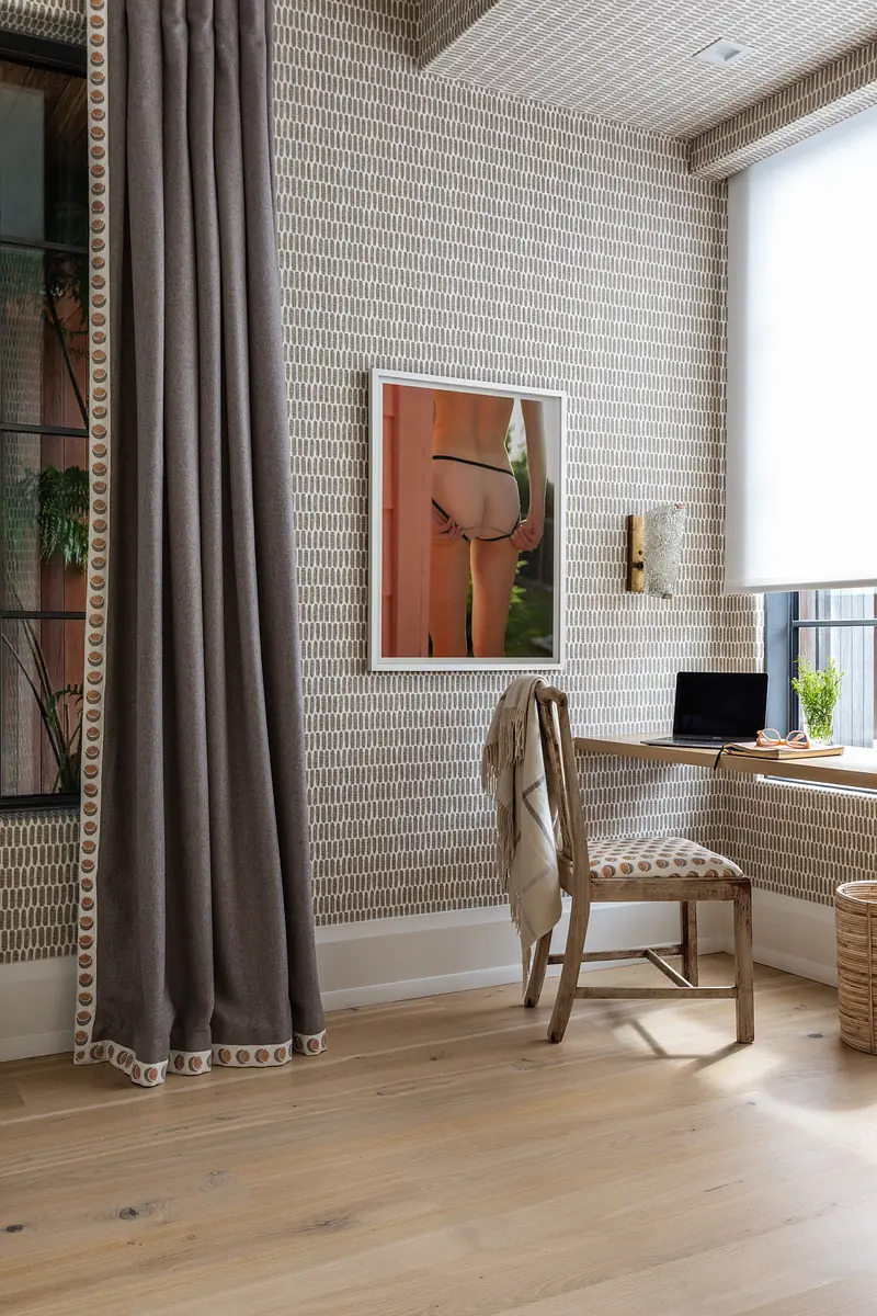 Small enclosed space with desk, laptop, chair, basket, patterned wallpaper, and large window with gray curtain.