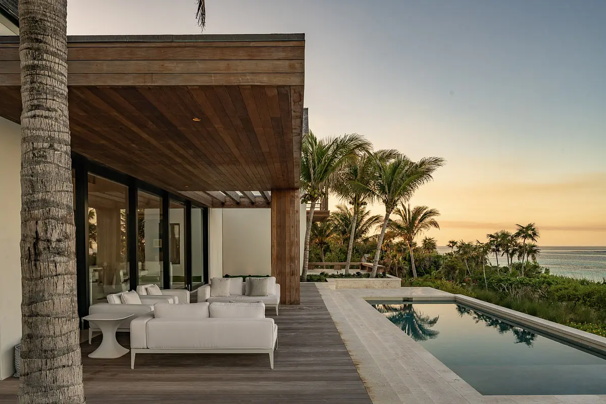 Outdoor patio with white lounge chairs, circular table, swimming pool, and palm trees at sunset.