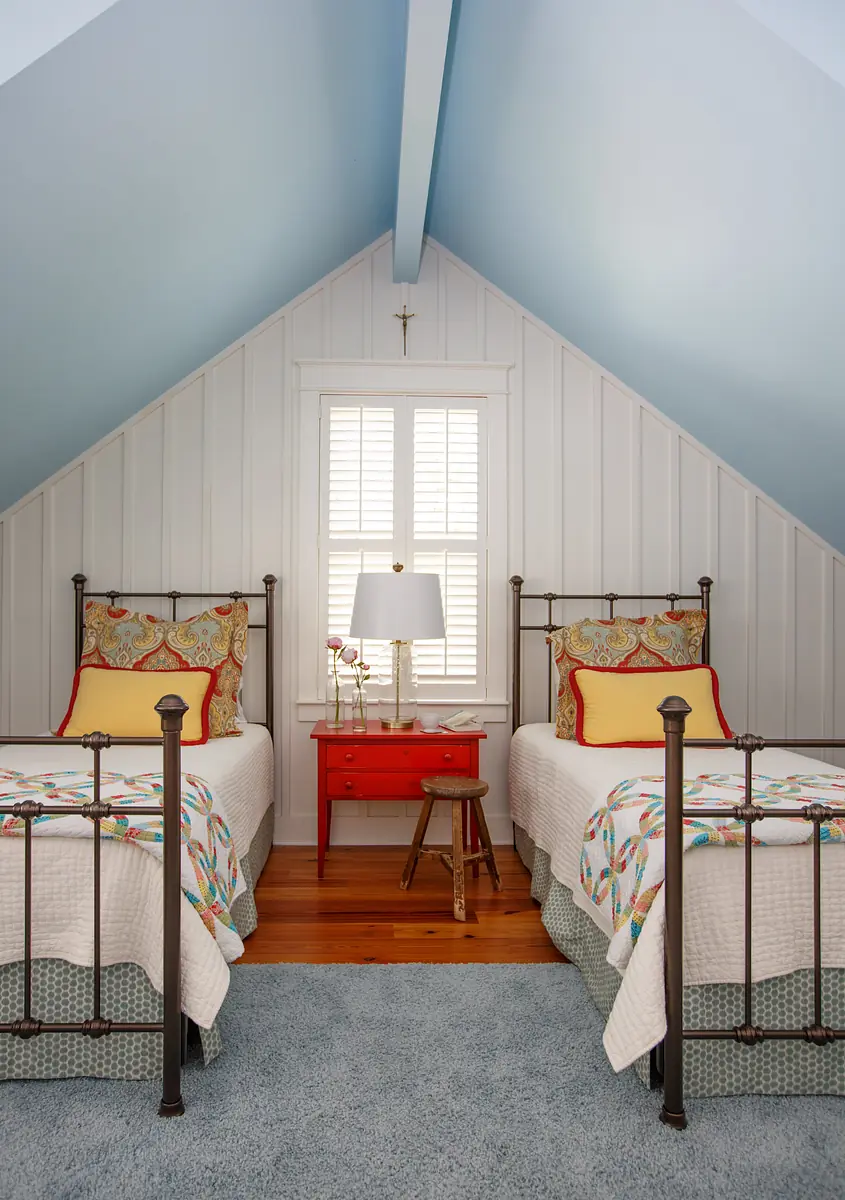 Bedroom with two metal frame beds, red nightstand, blue area rug, sloped ceiling, and window with shutters.