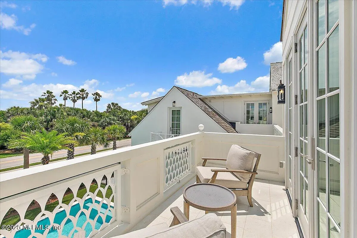 Deck with two light chairs, small table, lattice railing, palm trees, and a white building in the background.