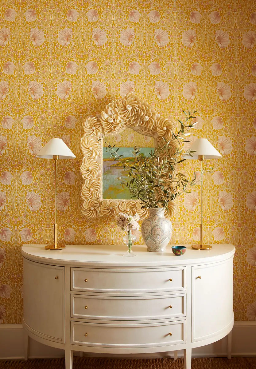Decorative wall with floral wallpaper, white sideboard, table lamps, mirror, and vase with greenery.