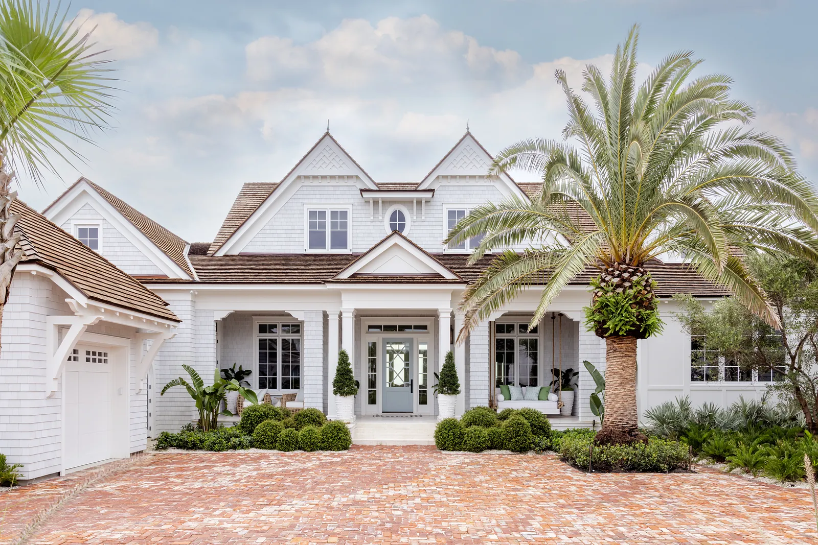 Front exterior featuring brick walkway, porch with columns, and potted plants amidst palm trees.