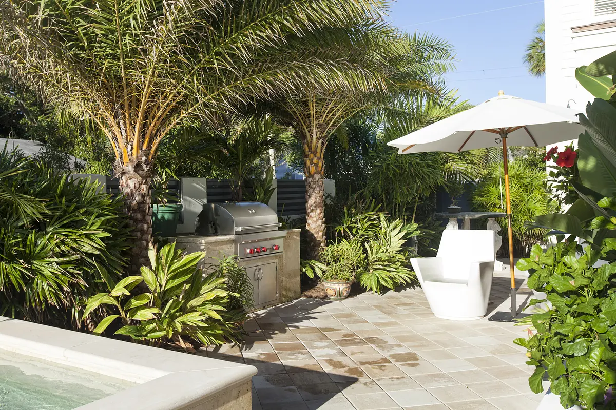 Patio area with white lounge chair, grill, large umbrella, and tropical plants on tiled flooring.