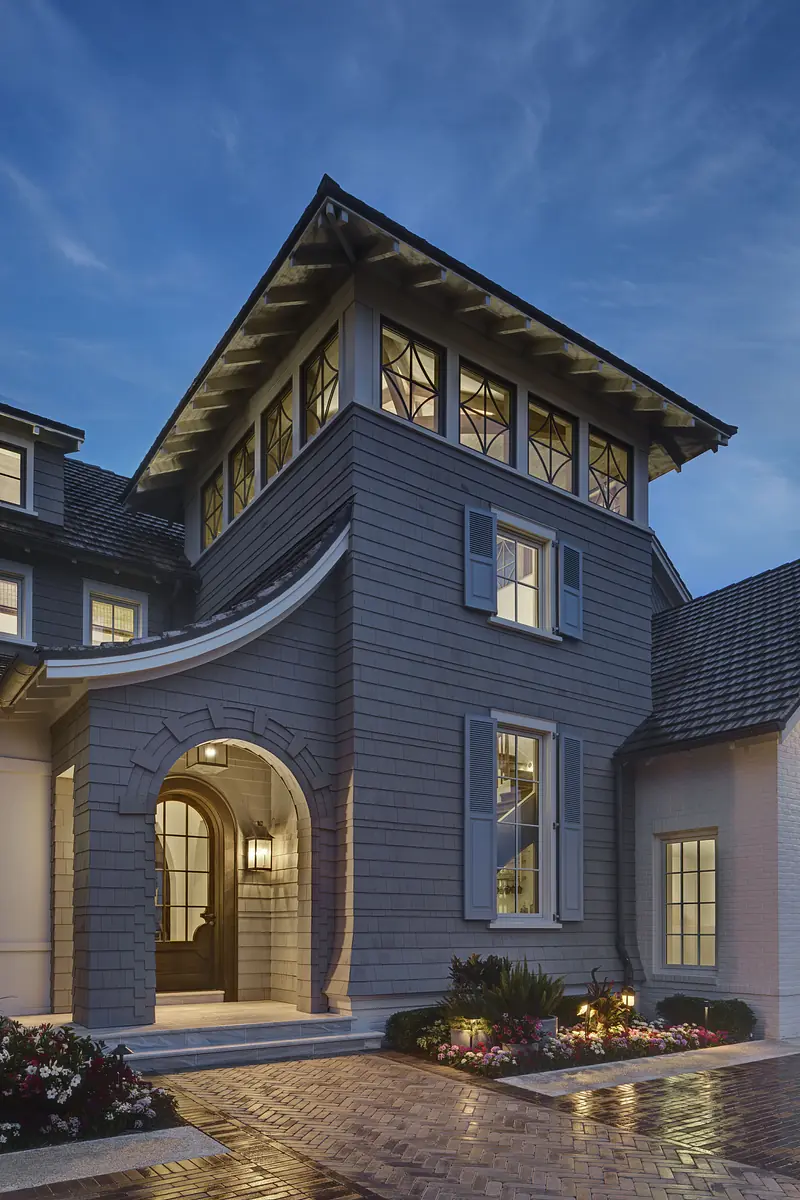 Exterior front of a house with gable roof, gray siding, blue shutters, arched entryway, and landscaped flower beds.