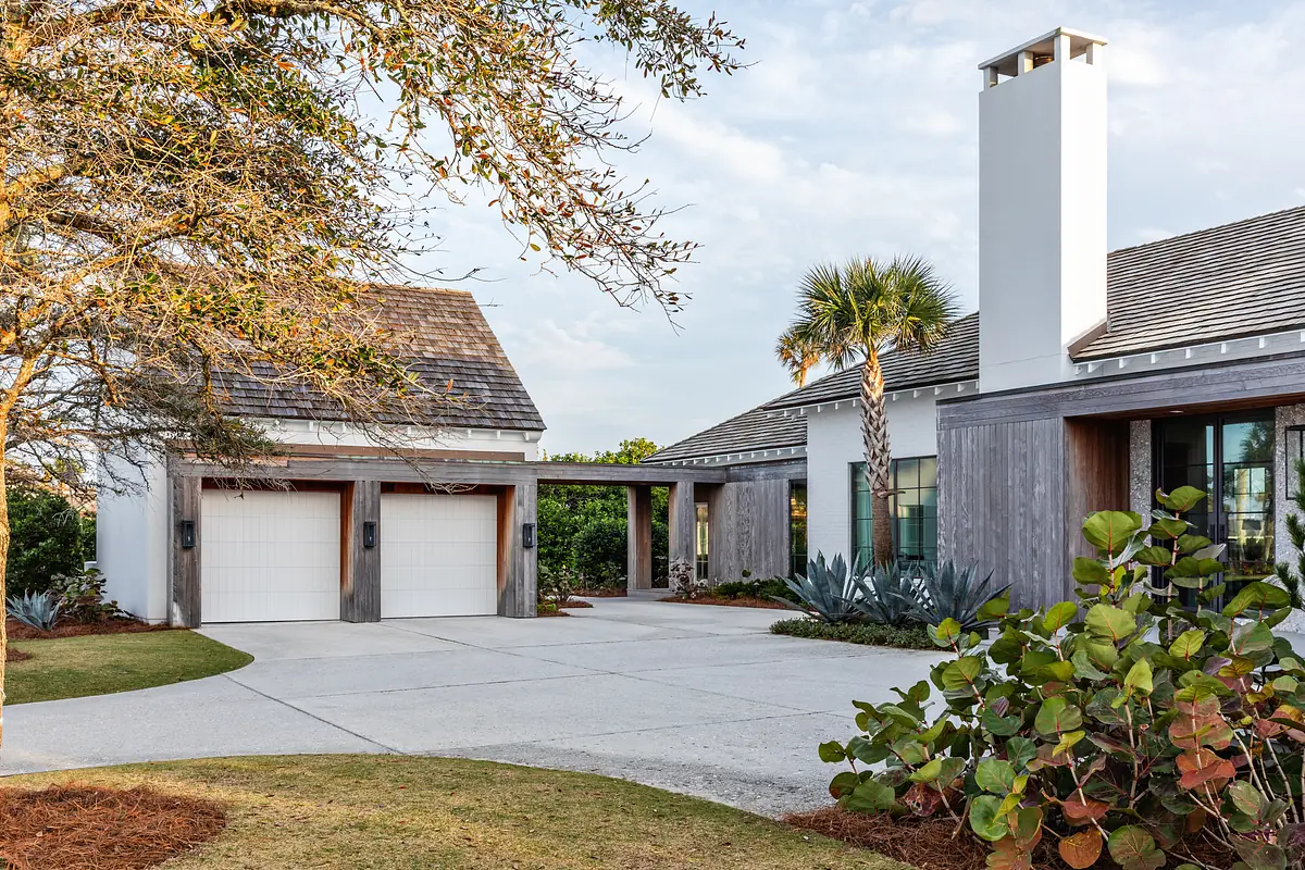 Front exterior featuring a garage, sloped roof, chimney, driveway, grass, shrubs, and palm trees.