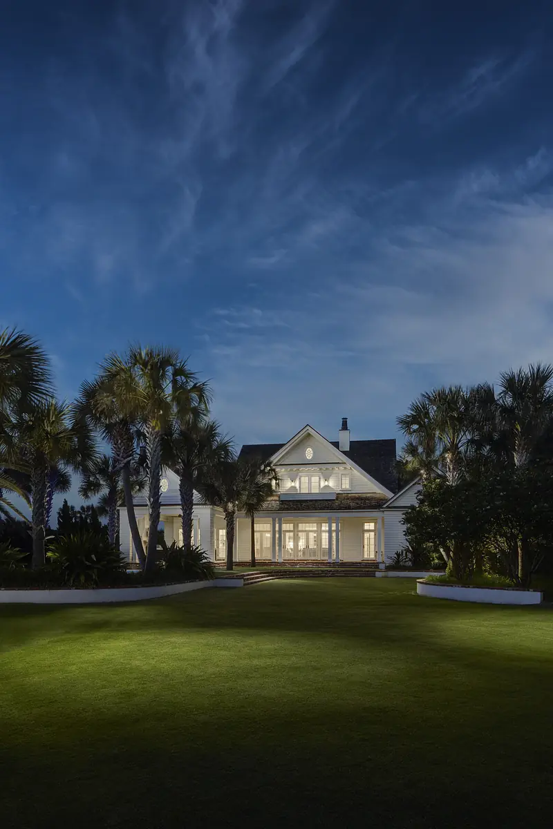 Front exterior of a two-story white house with large windows, porch, lawn, and palm trees at night.