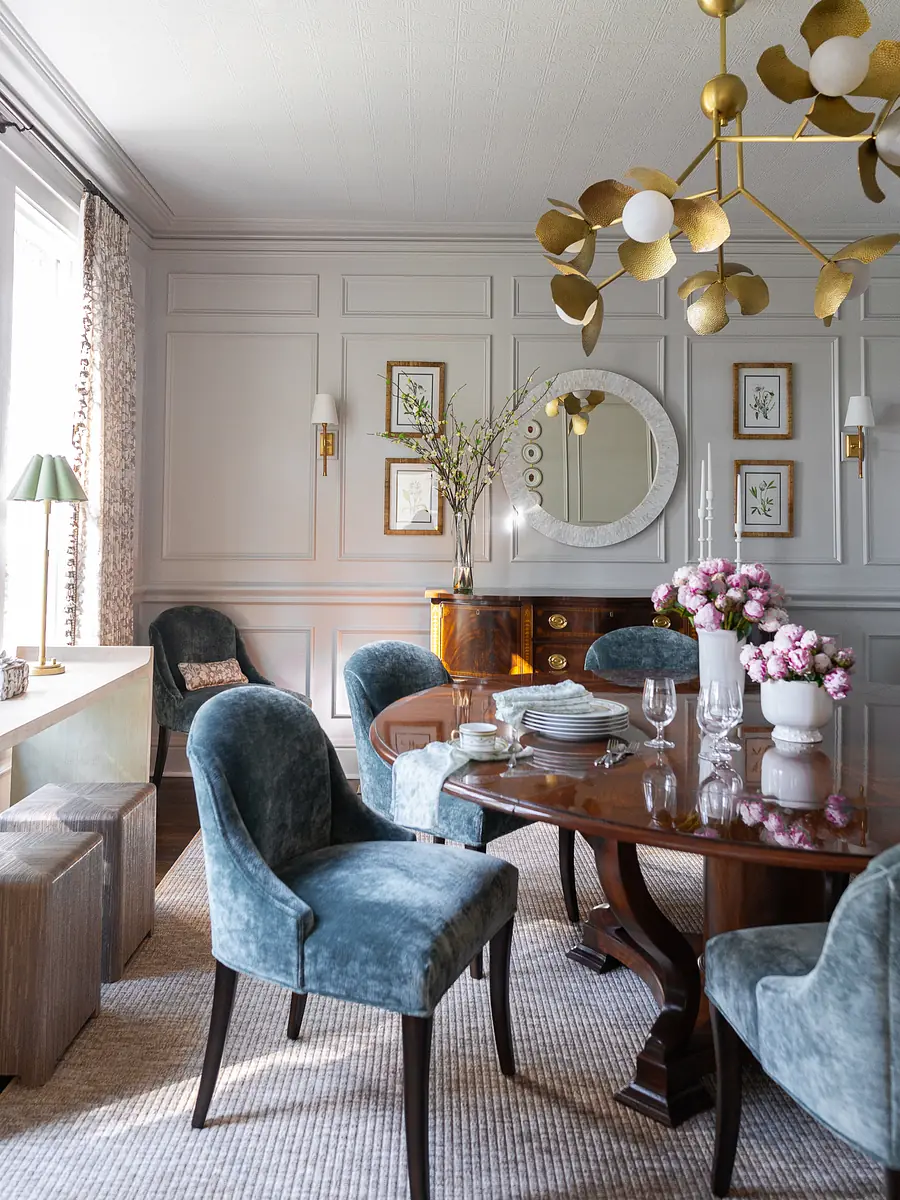 Dining room with round wooden table, teal chairs, paneled walls, mirror, sideboard, and floral decor.