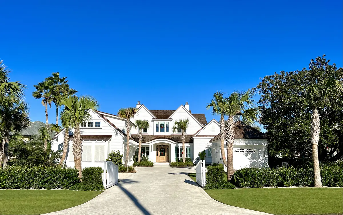 Front exterior of a house with white façade, brown shingles, central door, palm trees, and manicured lawn