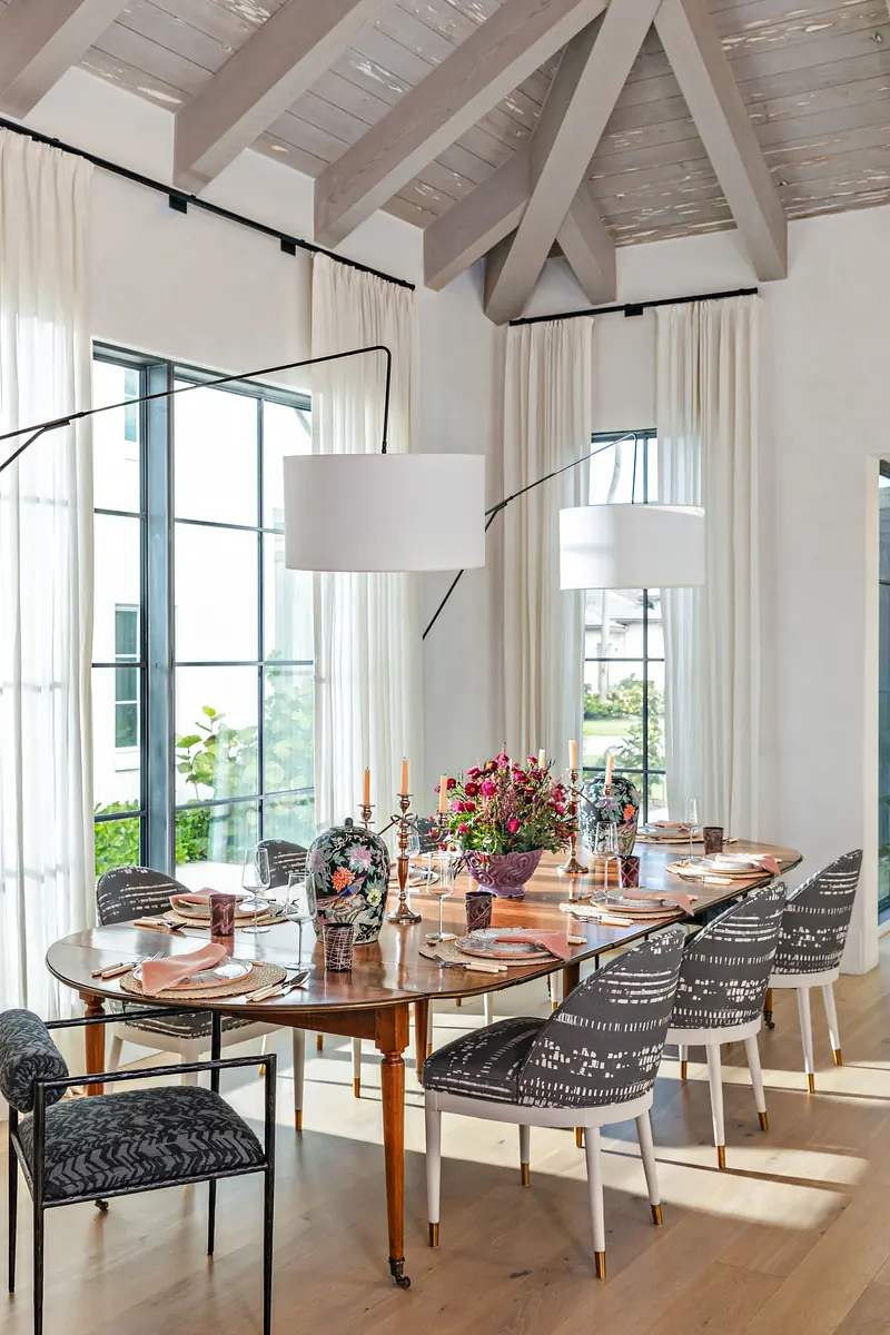 Dining room with round wooden table, patterned chairs, flowers, large windows, and pendant lights.