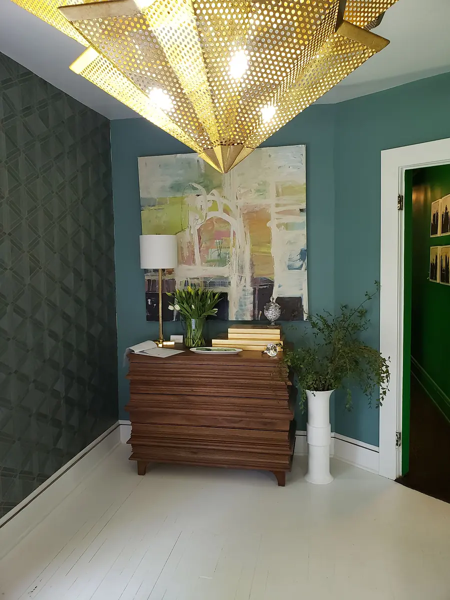 Foyer with green accent wall, wooden chest, abstract painting, lamp, vase with flowers, and potted plant.