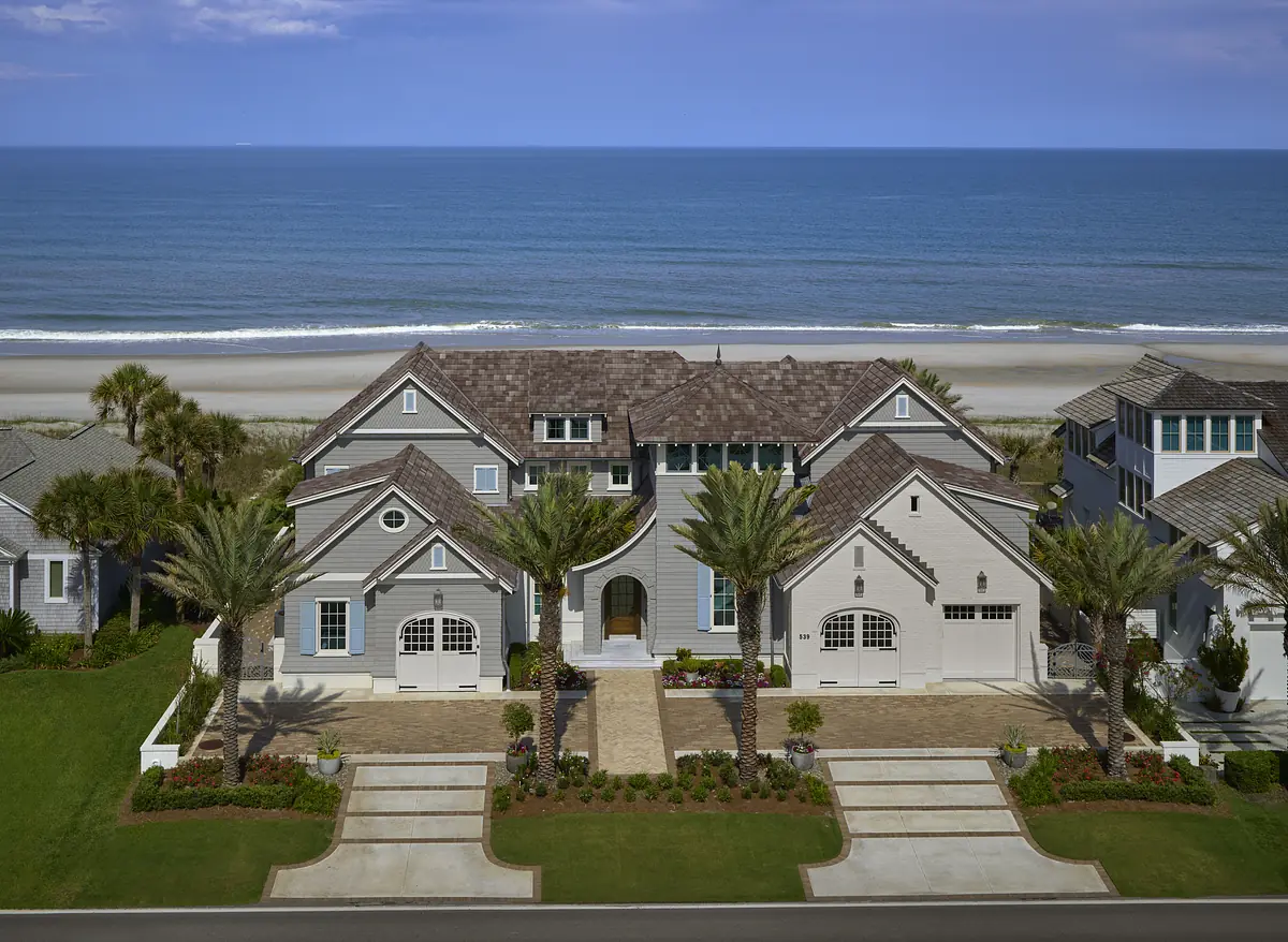 Front exterior view of a large home with gray siding, arches, large windows, palm trees, and brick paver driveway.