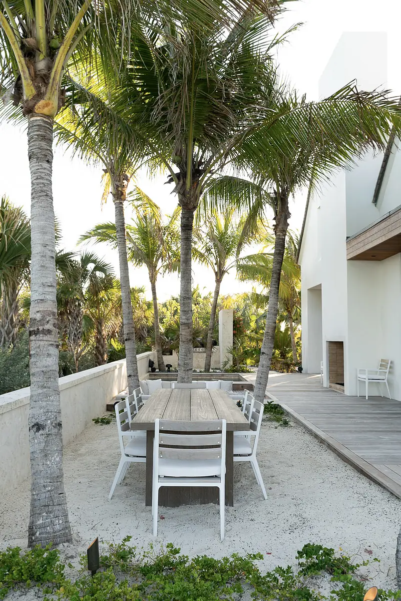Outdoor patio with wooden dining table, white chairs, palm trees, sandy ground, and wooden deck.
