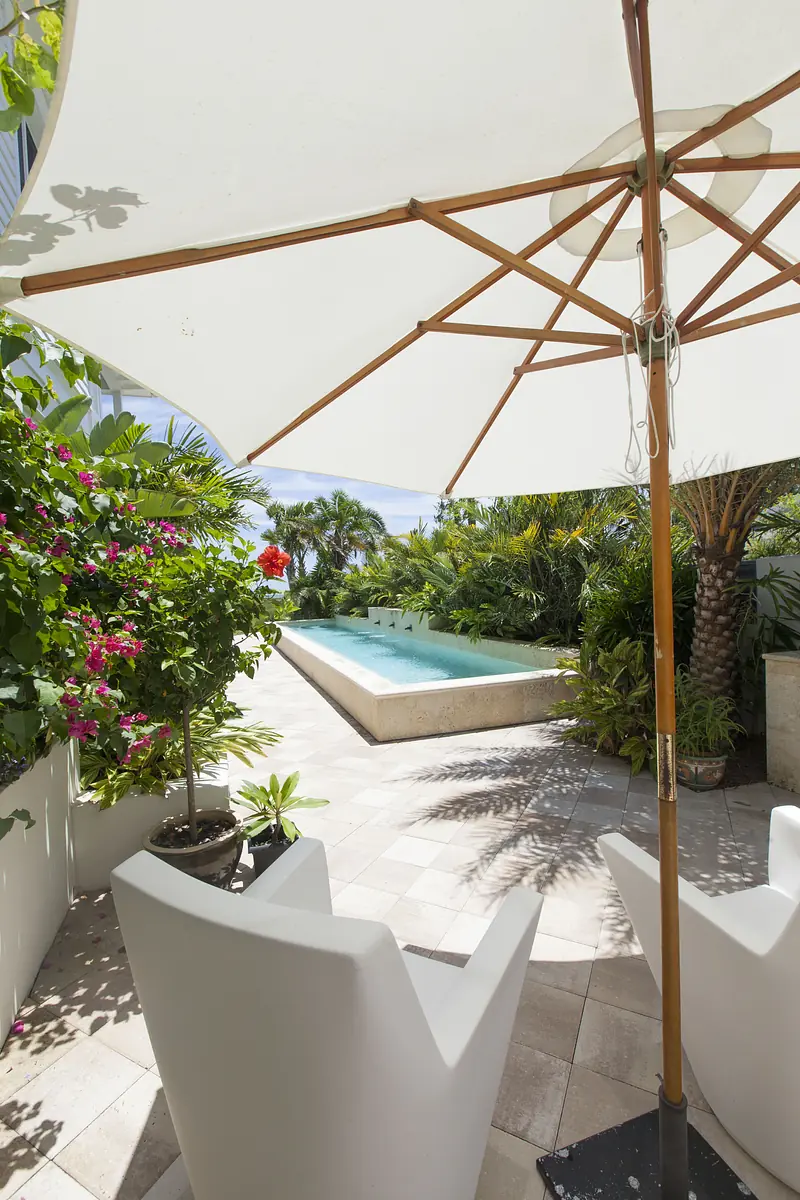 Patio with white chairs, large umbrella, tropical plants, and swimming pool in the background.