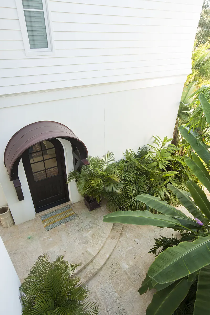 Exterior entrance with brown door, arched canopy, tropical plants, and stone pathway.