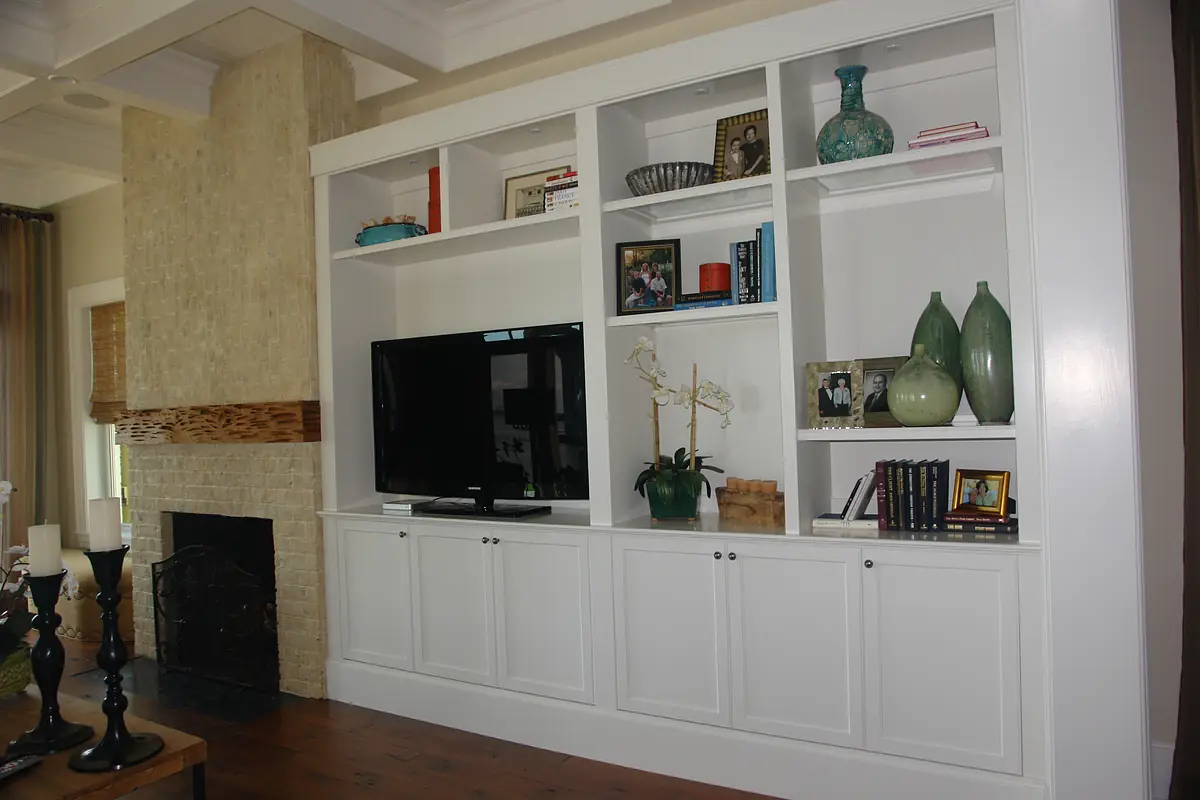 Living room with built-in cabinetry, television, green vases, books, brick fireplace, and hardwood flooring.