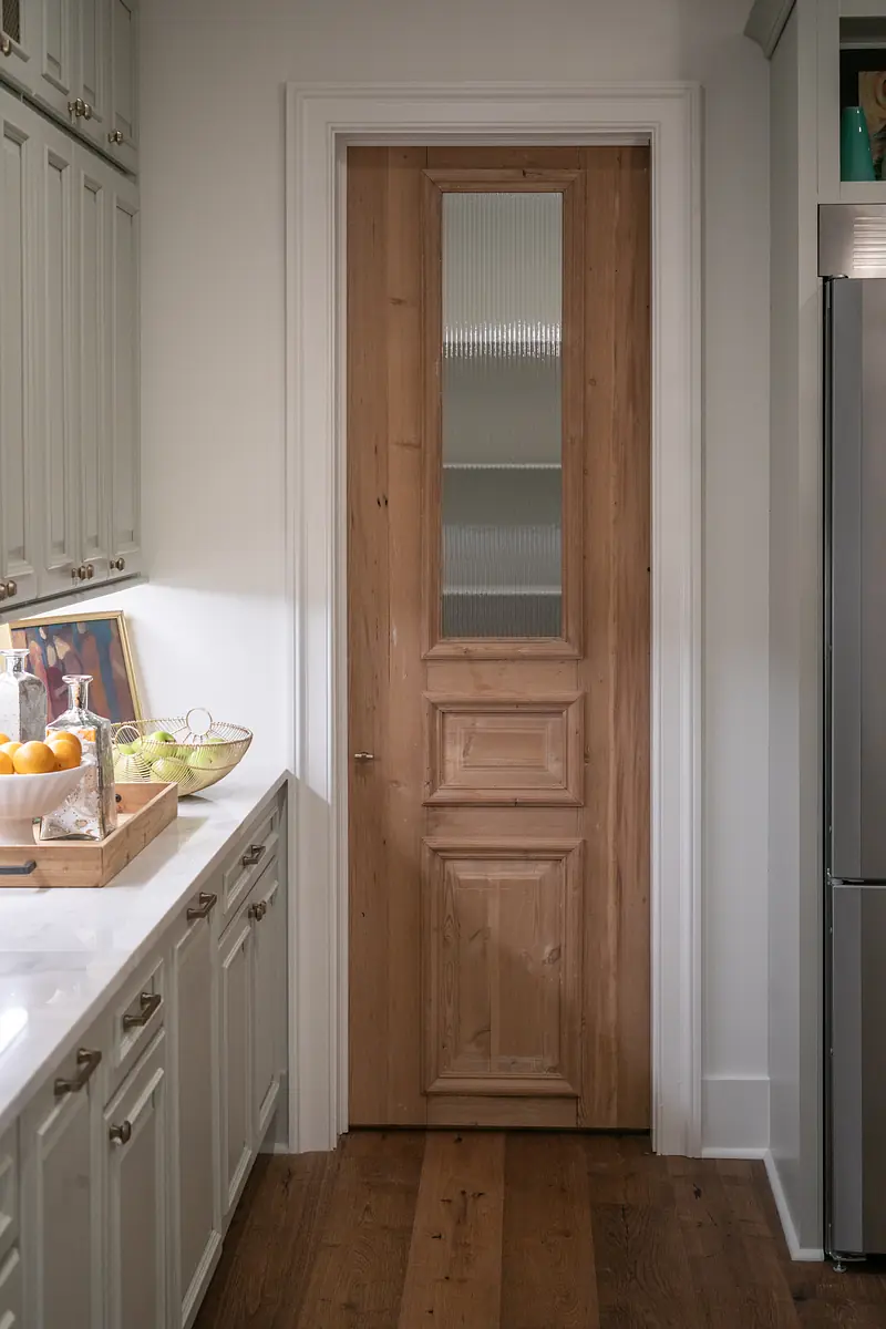 Kitchen with light green cabinetry, white countertop, wooden door, and fruit bowl.