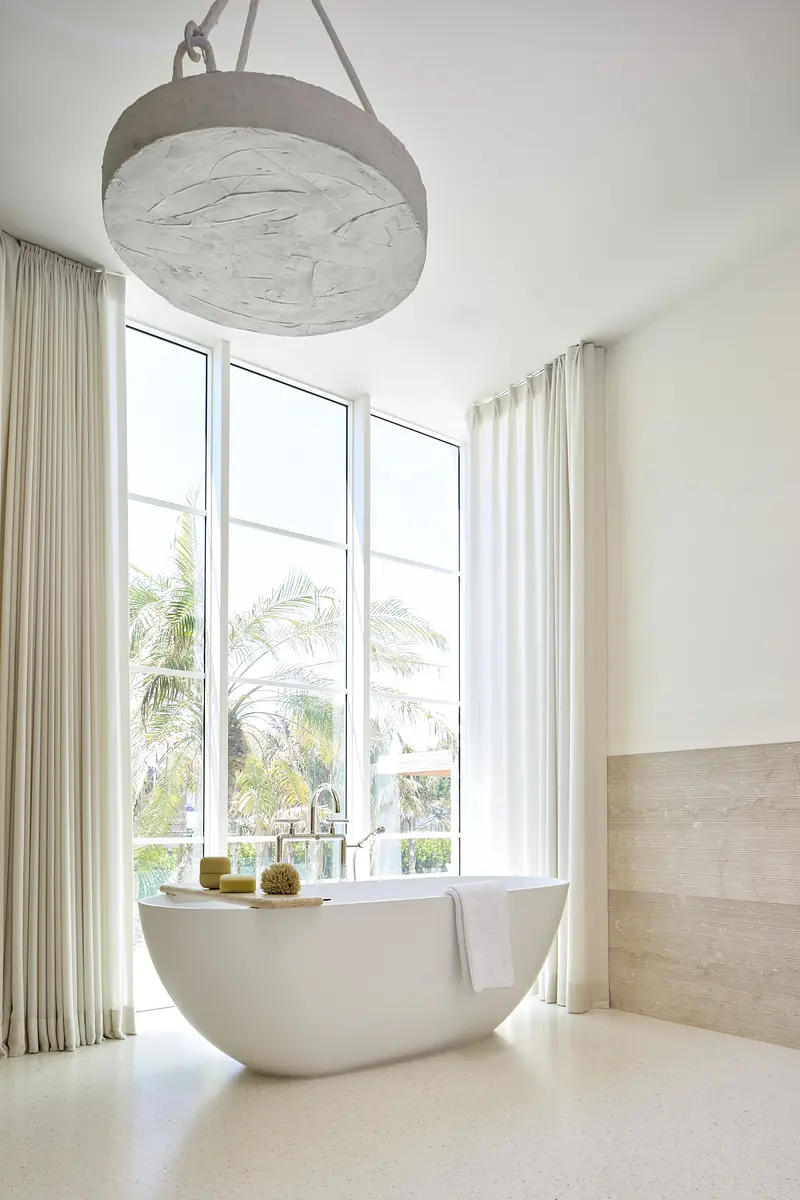 Bathroom with freestanding white tub, large windows, sheer curtains, and decorative items.