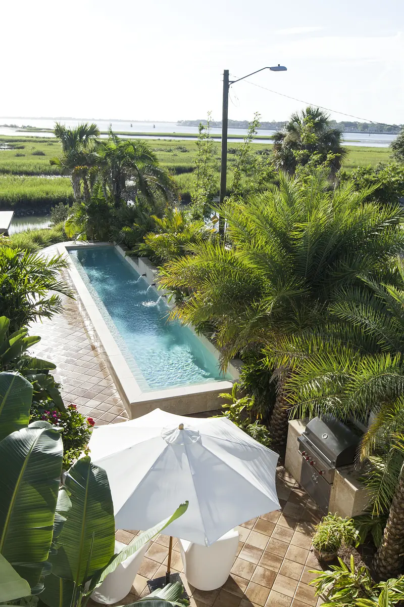 Outdoor deck with rectangular pool, tropical plants, umbrella, chair, and barbecue grill overlooking water.