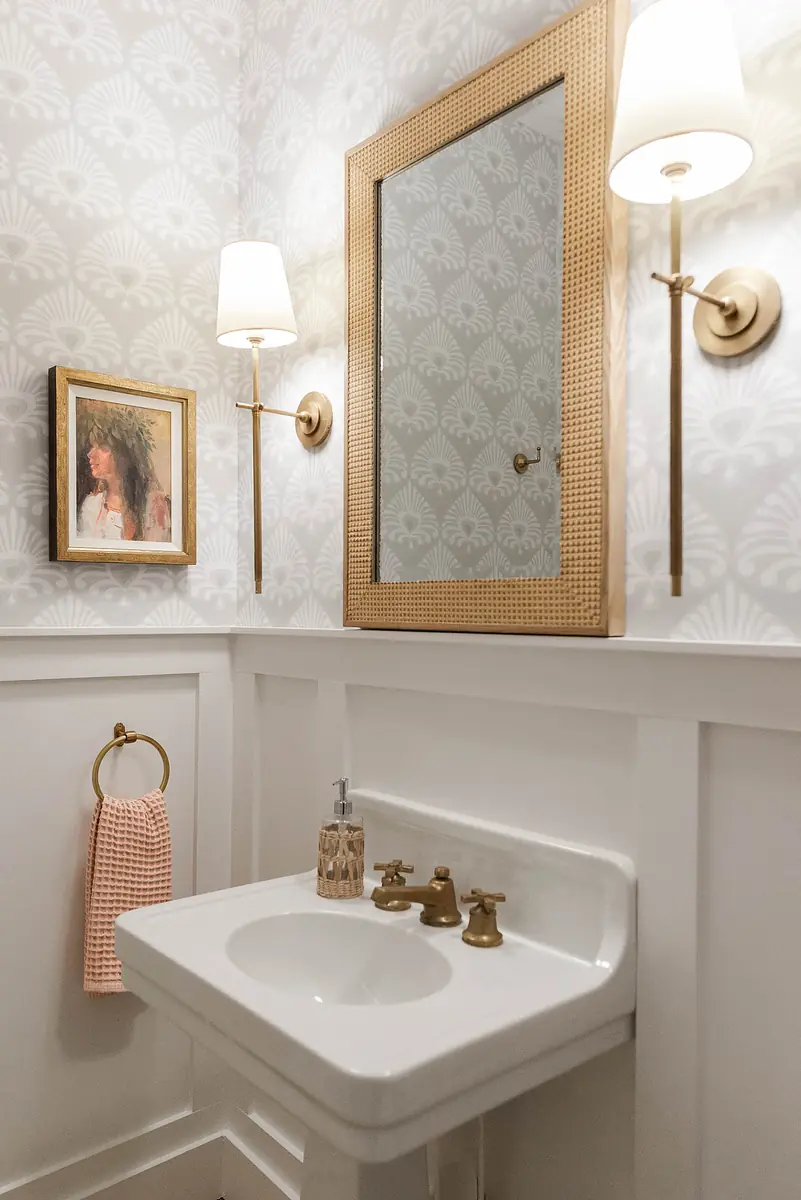 Powder room with wall-mounted sink, faucet, mirror, towel ring, sconces, patterned walls, and white wainscoting