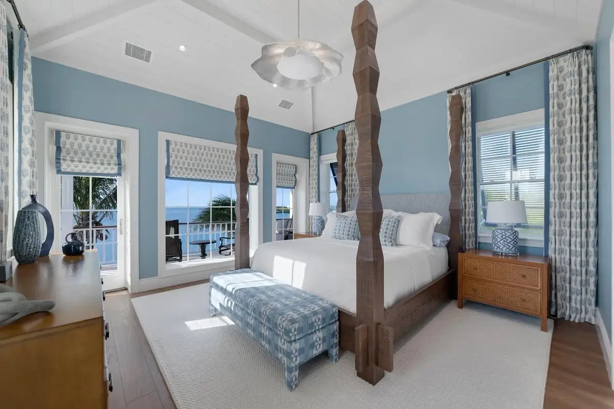 Bedroom with four-poster bed, light blue quilt, nightstands, lamps, large windows, and a dresser