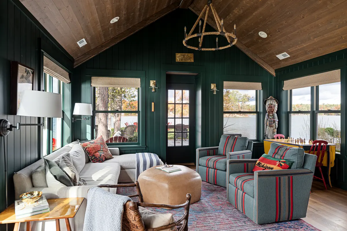 Living room with green paneled walls, gray sofa, striped armchairs, ottoman, and large windows providing outside views.
