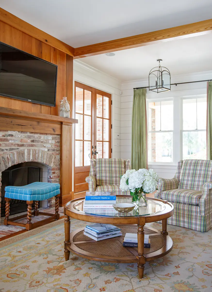 Living room with coffee table, plaid armchairs, brick fireplace, television, and large windows with curtains