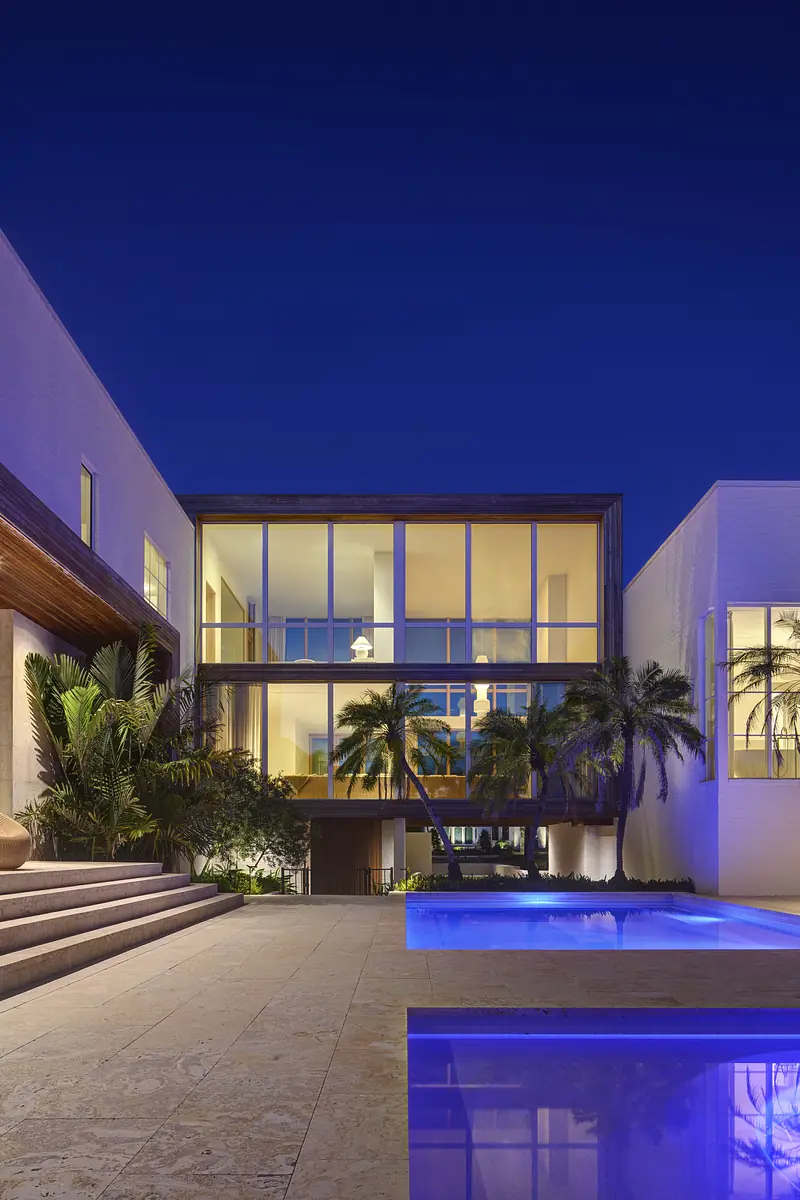 Exterior rear view of a modern house with large windows, pool, and tropical landscaping at night.